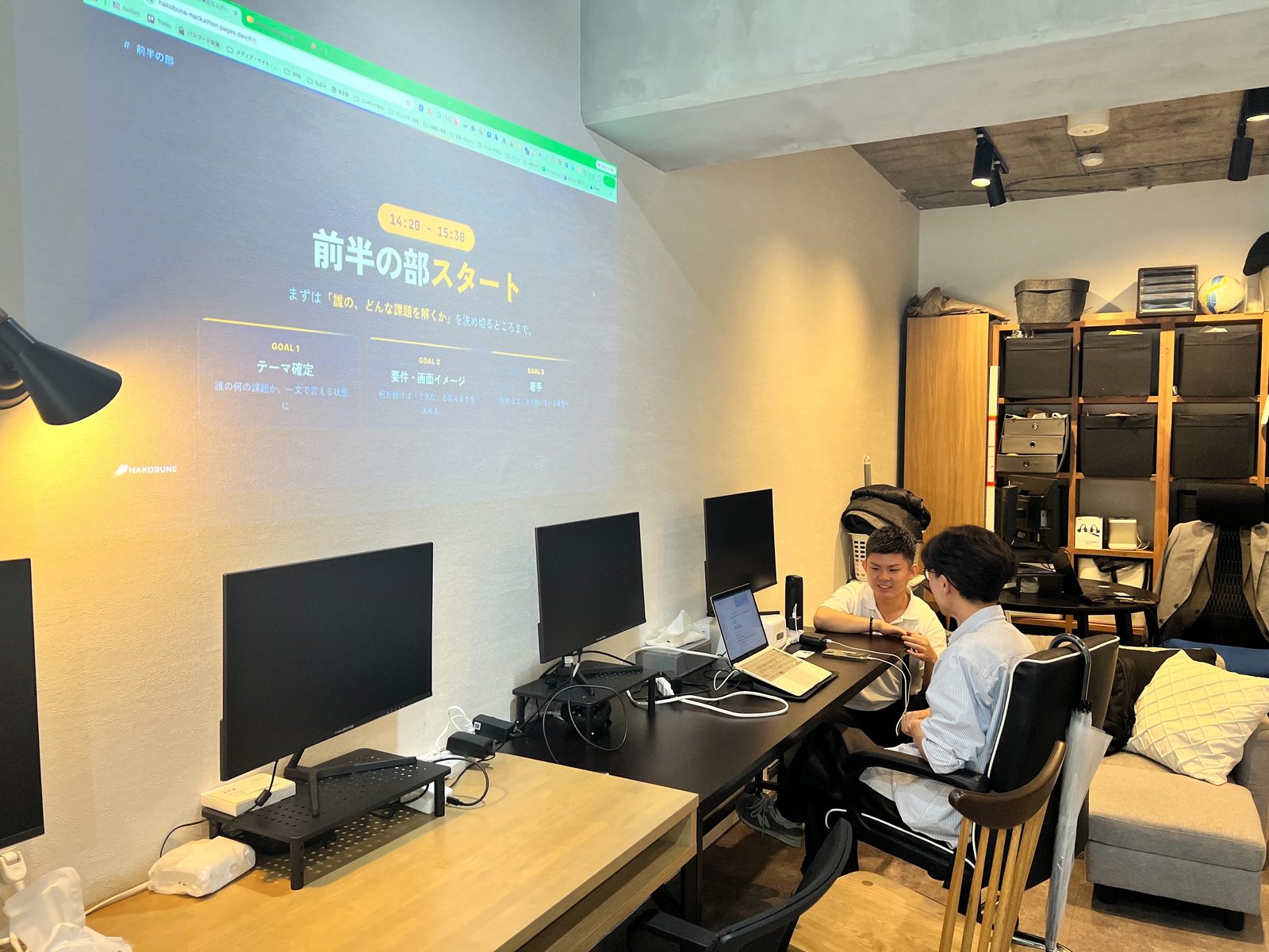 ハッカソン会場で、講師が参加者のノートPCを見ながら1対1でレクチャーしている様子。背面の壁には前半の部の進行スライドが投影されている